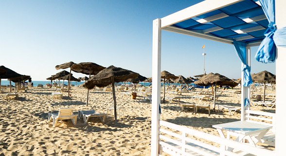 Beach with lots of sun loungers and thatched huts, including a white wooden pavilion with blue curtains.