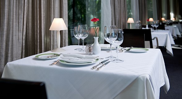 Elegant table in a stylish restaurant with white tablecloth, four wine glasses, cutlery and a red flower in a vase.