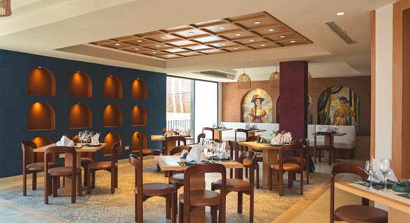 Modernes Restaurant Mit Tischen & Bestuhlung aus Hotel, in warmen Farben eingerichtet, Dunkelblaue Wand mit beleuchteten Regalen und Ornamenten an der Wand