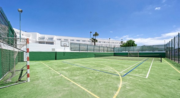 Multifunktions-Sportplatz mit grünem Bodenbelag, Toren, Basketballkorb und Tennisnetz vor weißem Hotelgebäude bei Sonnenschein.