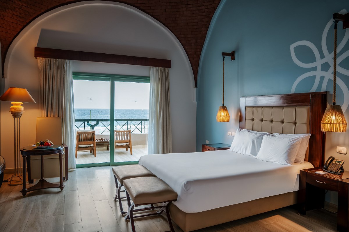 Großzügiges Hotelzimmer mit gewölbter Decke, Doppelbett mit roten Akzenten, Sitzbereich und Balkon mit Ausblick auf die Stadt.