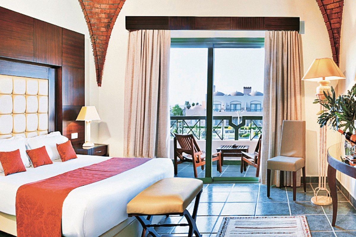 Stilvolles Hotelzimmer mit Doppelbett, blauen Akzenten, gewölbter Backsteindecke und Balkon mit Blick auf eine Resortanlage.