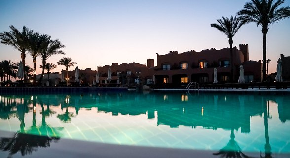 Beleuchteter Hotelpool bei Sonnenuntergang, mit Palmen und flachen Gebäuden im Hintergrund, stimmungsvoll gespiegelt im Wasser.