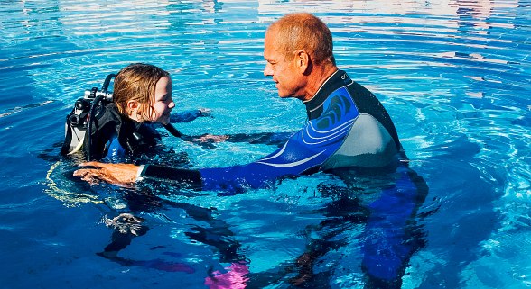 Tauchlehrer im Neoprenanzug unterrichtet ein Kind mit Tauchausrüstung im klaren Wasser eines Pools.