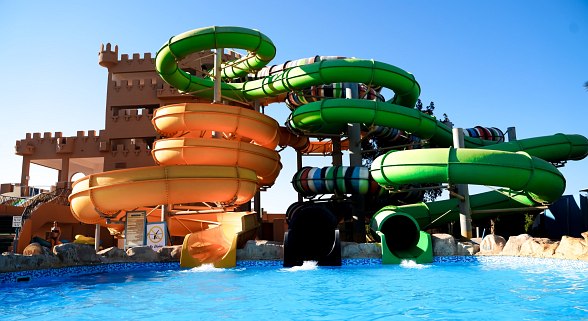 Wasserpark mit bunter Rutschenanlage vor einem burgähnlichen Gebäude, mehrere Rutschen münden in ein großes Schwimmbecken.