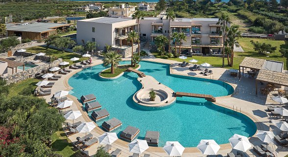 Großer Pool mit Insel, Palmen und Brücke, Liegen mit weißen Schirmen, Hotelgebäude im Hintergrund, sonniger Tag.