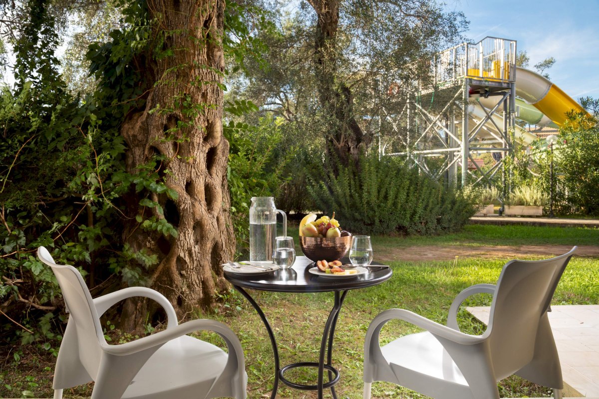 „Terrasse mit Tisch und Stühlen, Blick auf Garten und moderne Gebäude, serviertes Frühstück bei Sonnenschein.