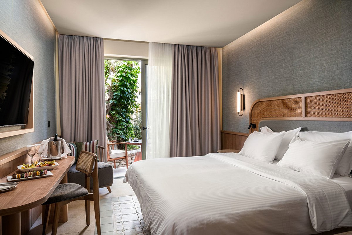 „Modernes Hotelzimmer mit Doppelbett, Holzdetails, Schreibtisch und großem Fenster mit lichtdurchlässigem Vorhang.