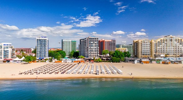  Hotel Sentido Bellevue Beach am Strand mit vielen Liegestühlen und Hotels im Hintergrund unter blauem Himmel. Modernes Hotelzimmer mit zwei Betten, blauem Sessel, TV und Balkon mit Meerblick.