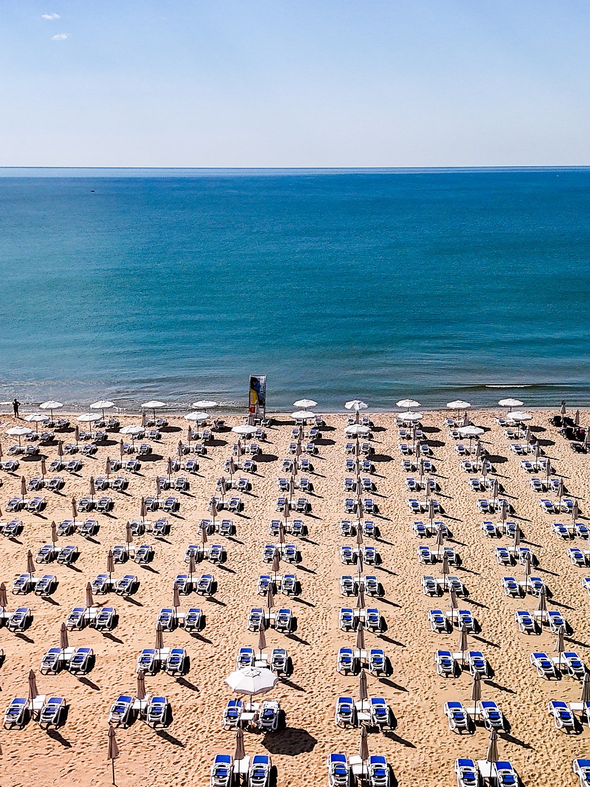 Weitläufiger Strand mit strahlendblauem Wasser, feinsandig und viele Sonnenschirme mit Sonnenliegen.