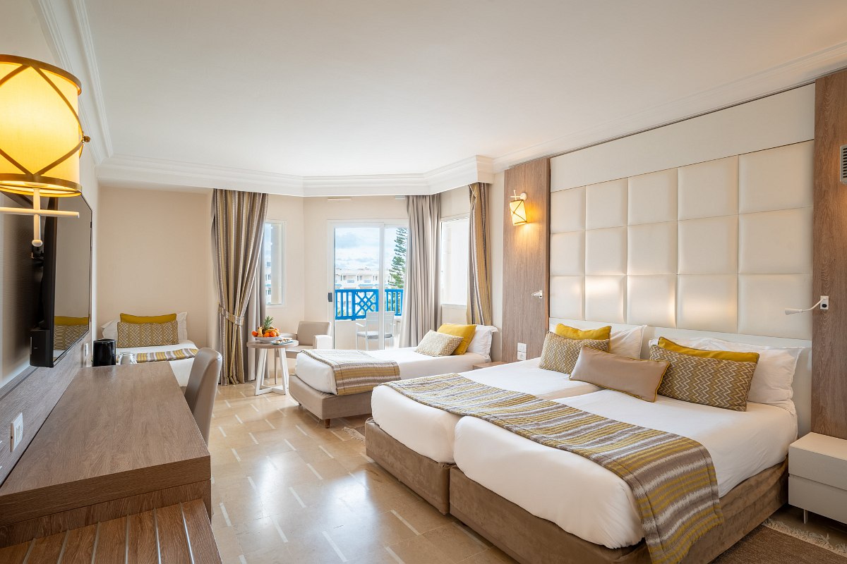 Modernes Hotelzimmer mit großem Bett, zusätzlicher Liege, Balkon mit Meerblick und warmen Gelb-Braun-Tönen.