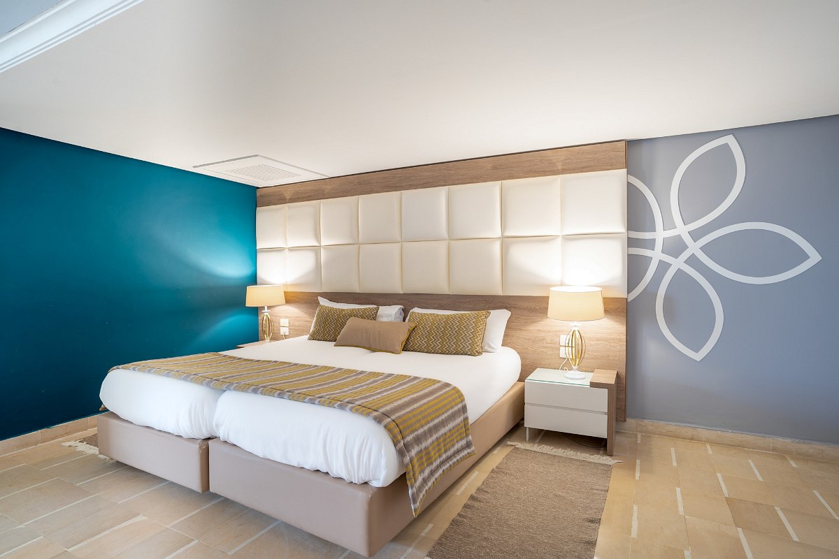 Helles Hotelzimmer mit Doppelbett, zusätzlichem Bett, Balkon mit Meerblick, Schreibtisch und Flachbild-TV.