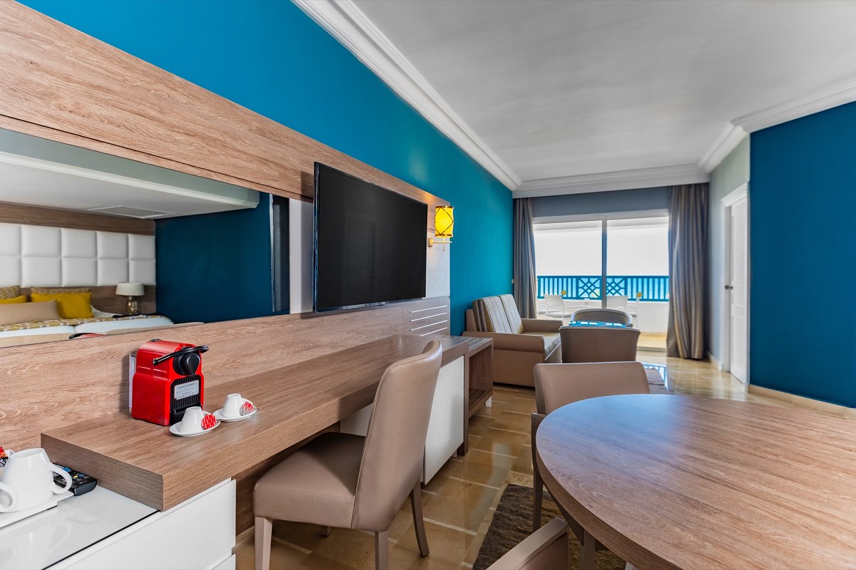 Modernes Hotelzimmer mit großem Bett, zusätzlicher Liege, Balkon mit Meerblick und warmen Gelb-Braun-Tönen.