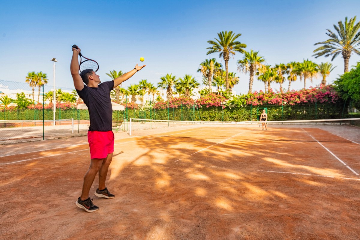 Mann beim Tennisaufschlag auf einem Sandplatz, umgeben von Palmen und Blumen unter klarem, blauem Himmel.