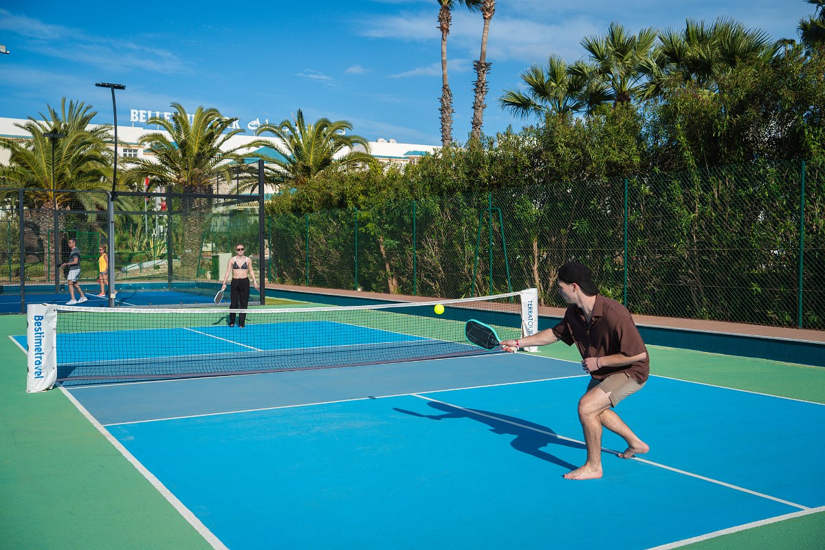 Mann beim Tennisaufschlag auf einem Sandplatz, umgeben von Palmen und Blumen unter klarem, blauem Himmel.