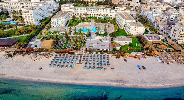 Luftaufnahme eines großen Strandhotels mit Poolanlage, Sonnenschirmen, Liegen und direktem Zugang zum Sandstrand.