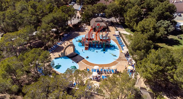 Wasserpark mit Piratenschiff-Spielplatz in flachem Pool, umgeben von Liegestühlen und Pinienwald aus der Vogelperspektive.