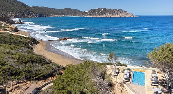 Küste mit Sandstrand, Wellen und Infinity-Pool mit Meerblick, umgeben von Pinien und felsiger Landschaft.