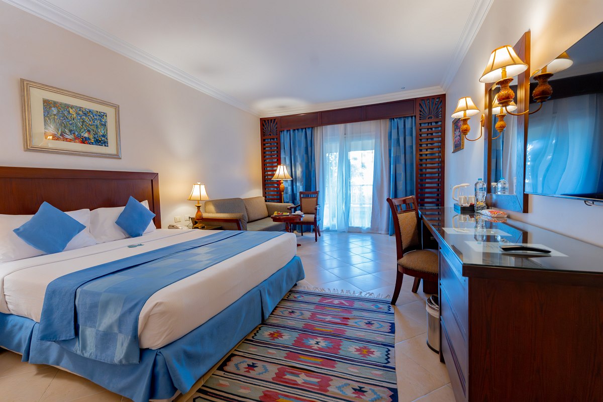 Helles Hotelzimmer mit Doppelbett, Sofa, Schreibtisch und Balkon, warme Beleuchtung und Blick ins Grüne durch große Tür.