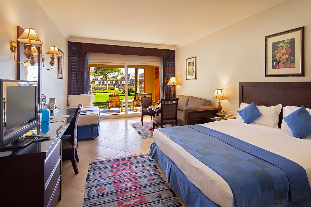 Helles Hotelzimmer mit Doppelbett, Sofa, Schreibtisch und Balkon, warme Beleuchtung und Blick ins Grüne durch große Tür.