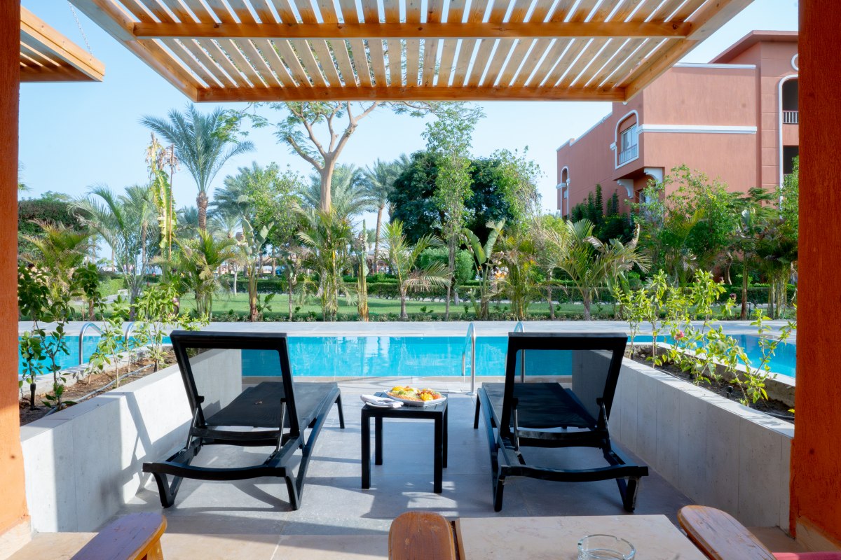Zwei schwarze Liegestühle mit einem kleinen Tisch dazwischen auf einer Terrasse mit Blick auf einen Pool, umgeben von Palmen und grünen Pflanzen.