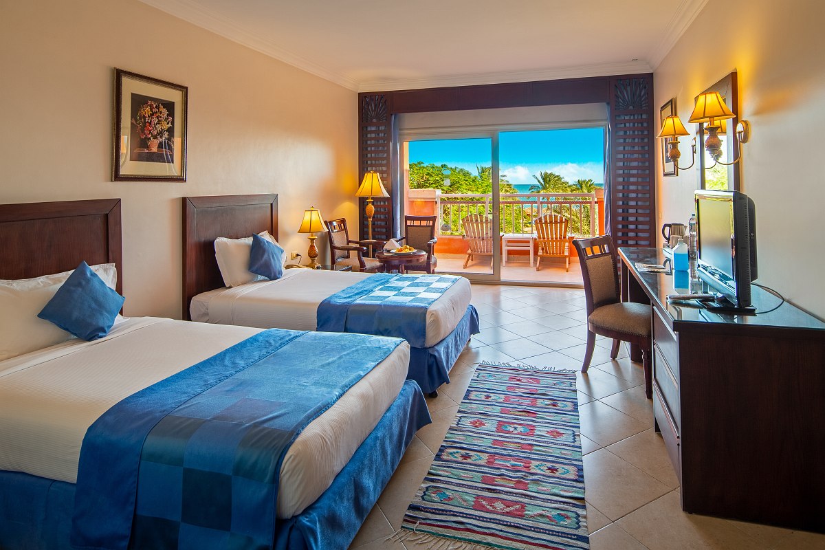 Hotelzimmer mit Doppelbett, Sofa, Schreibtisch und Balkon mit Sitzmöbeln, helle Fliesen und Blick ins Grüne bei Tageslicht.