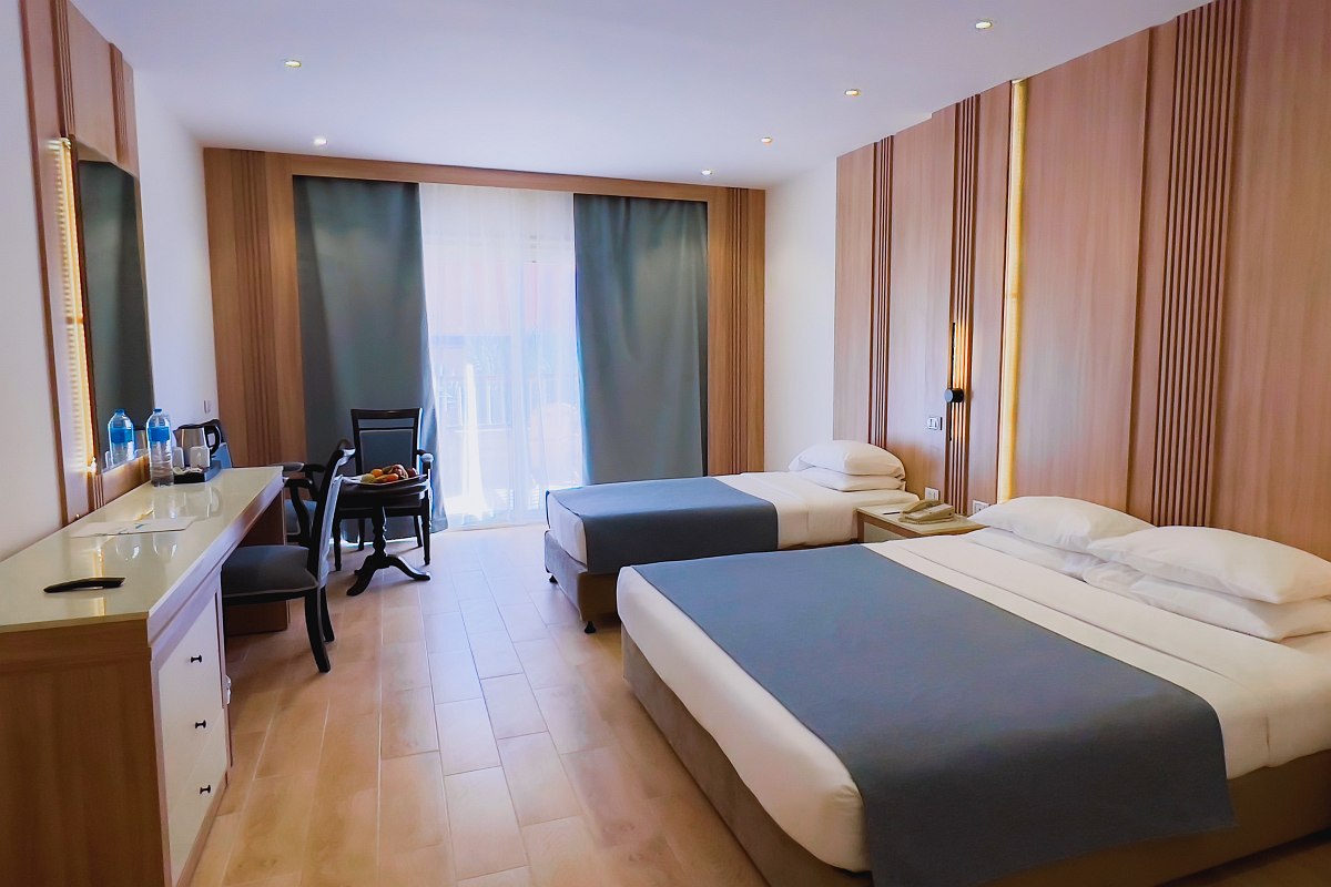 Helles Hotelzimmer mit Doppelbett, Sofa, Schreibtisch und Balkon, warme Beleuchtung und Blick ins Grüne durch große Tür.