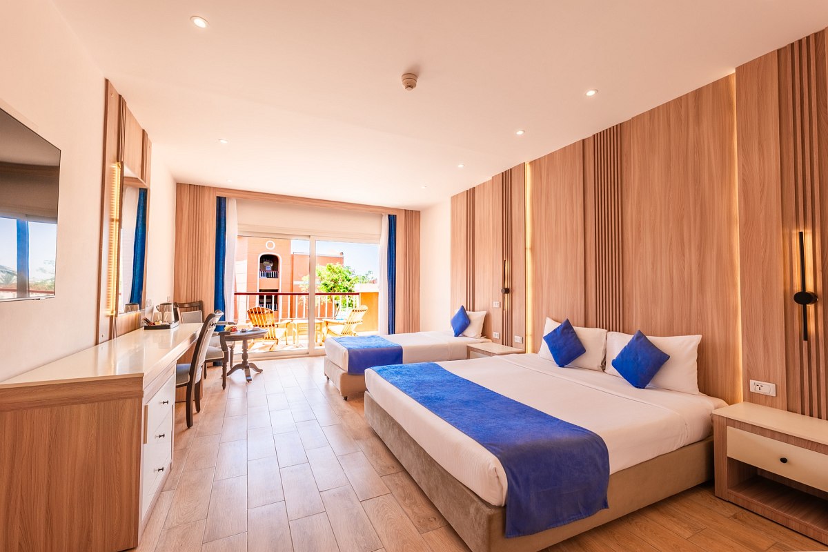 Großes, helles Hotelzimmer mit Doppel- und Einzelbett, Holzboden, blauen Akzenten und Balkon mit Blick auf grüne Umgebung.