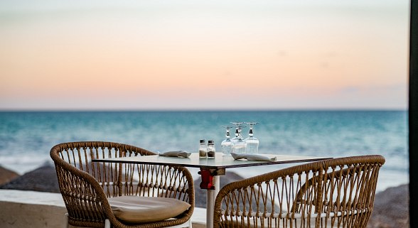 Gedeckter Tisch mit zwei Stühlen am Meer bei Sonnenuntergang mit Blick auf das ruhige Wasser.