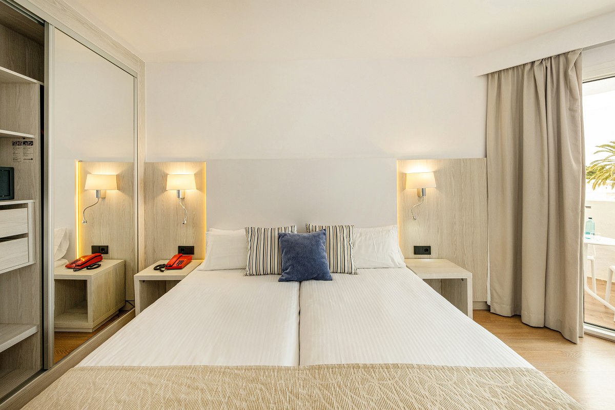 Helles Hotelzimmer mit Doppelbett, Schreibtisch, Balkonzugang und Blick auf den Himmel durch Glastür.