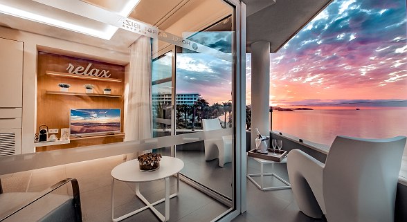 Modernes Hotelzimmer mit Balkon und Meerblick bei Sonnenuntergang, eingerichtet mit Lounge-Stühlen und Champagner-Service.