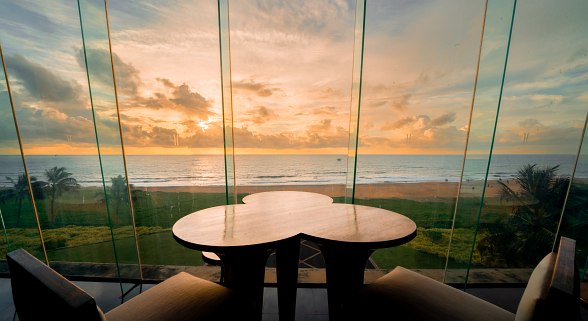 Stimmungsvoller Sitzbereich mit Glasfront und einzigartigem Tischdesign, Blick auf Strand und Meer bei Sonnenuntergang.