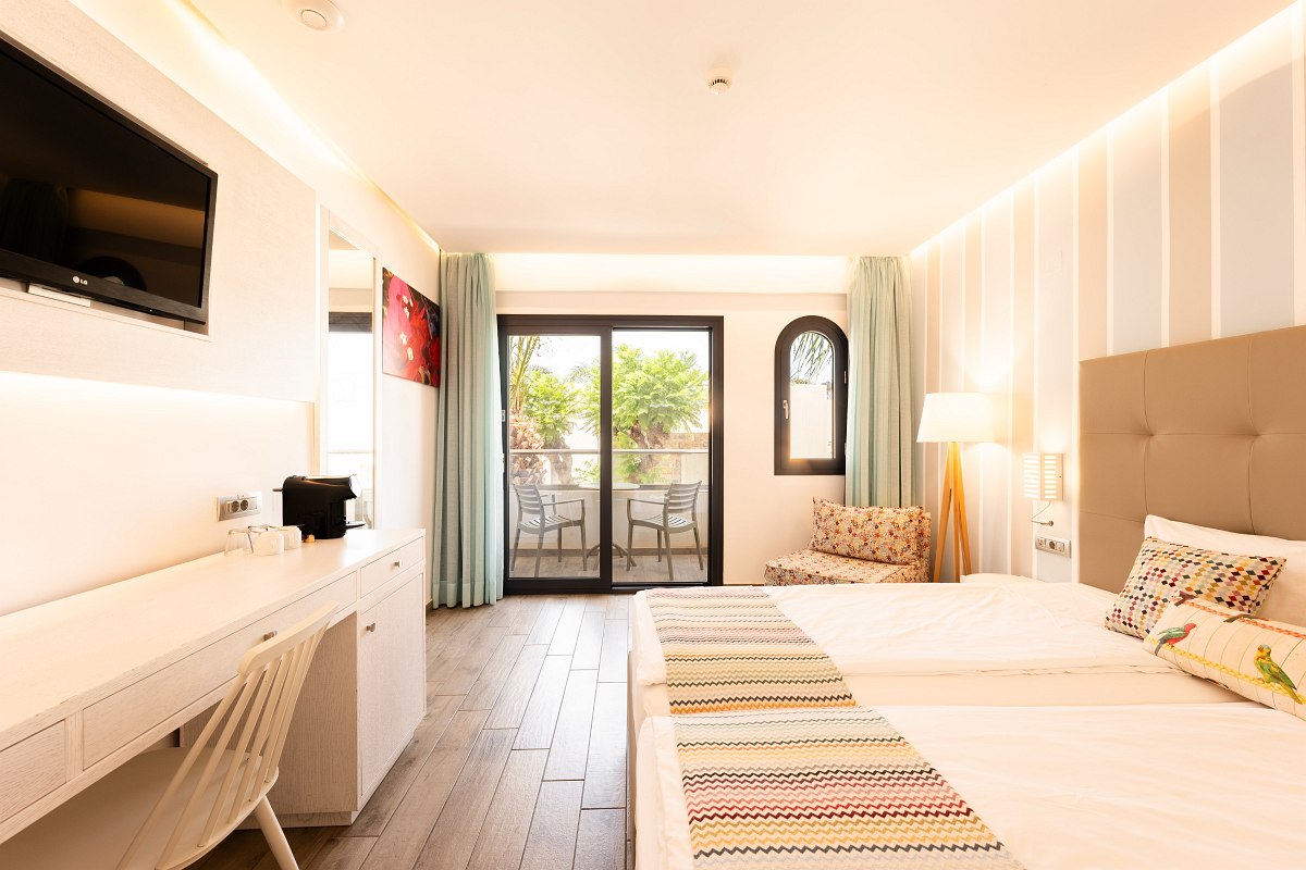 Helles Hotelzimmer mit Doppelbett, Balkon, Schreibtisch, Fernseher, Kaffeemaschine und gemütlichem Sessel.