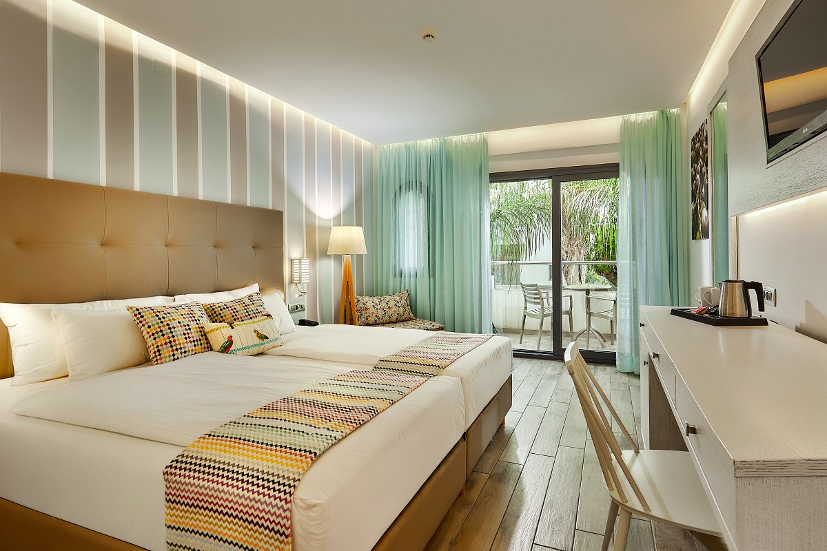 Helles Hotelzimmer mit Doppelbett, gemusterten Kissen, Schreibtisch und Balkon mit Blick auf grüne Bäume.
