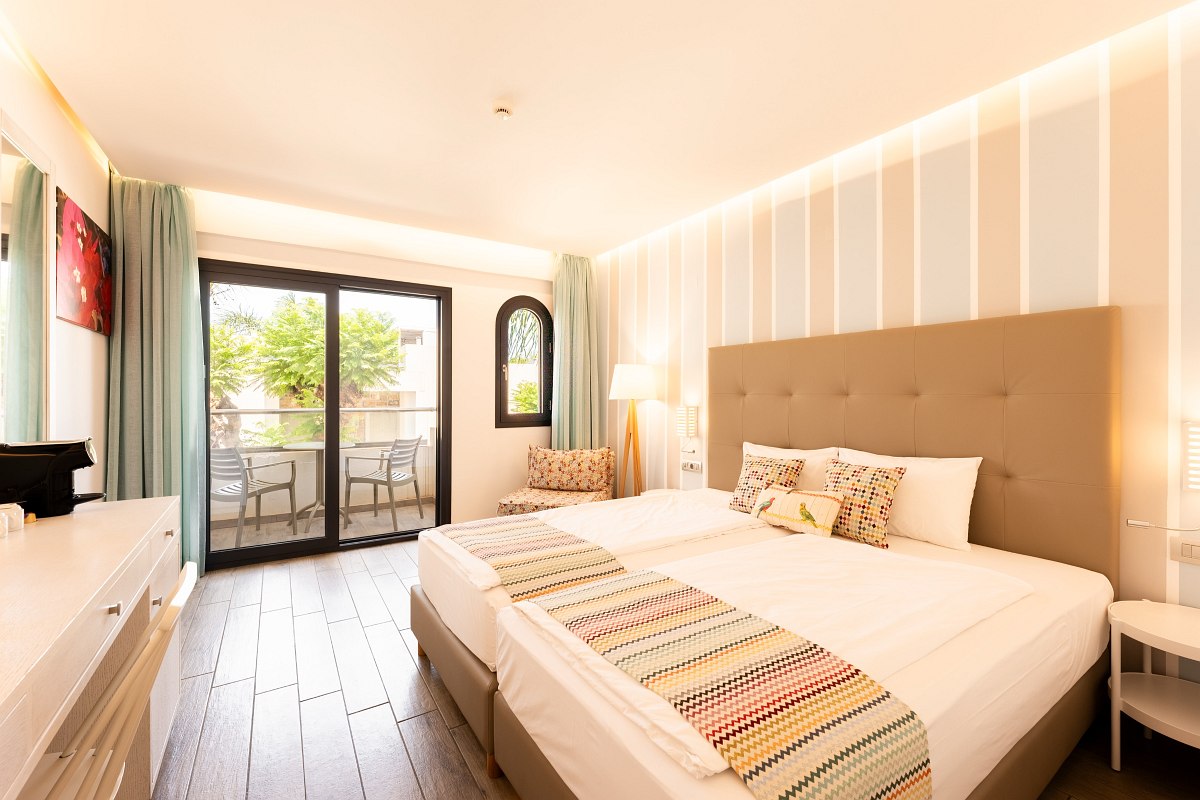Helles Hotelzimmer mit Doppelbett, gemusterten Kissen, Schreibtisch und Balkon mit Blick auf grüne Bäume.