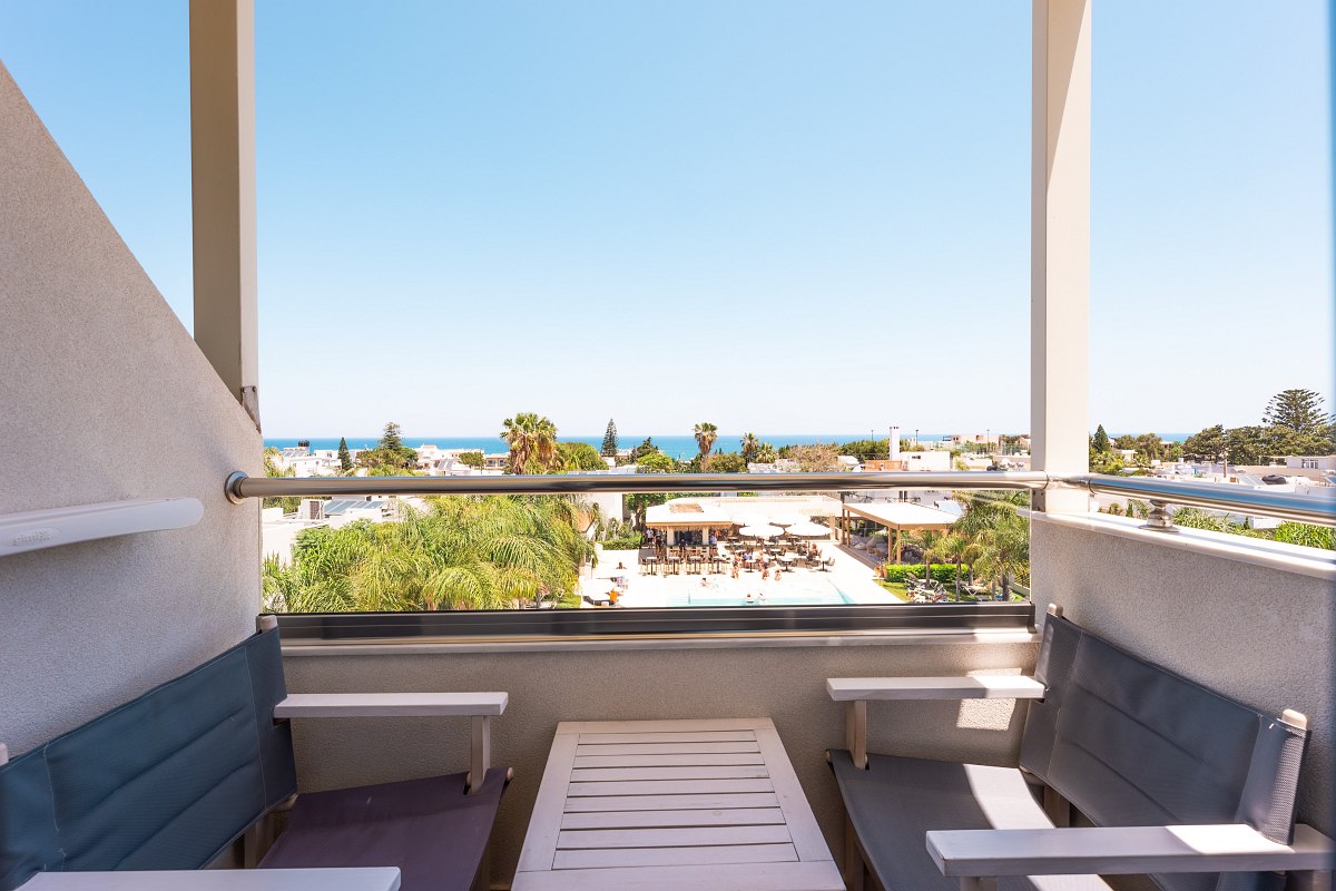 Kleiner Balkon mit zwei Sitzbänken und Tisch, Blick auf Poolbereich, Palmen und das Meer im Hintergrund.