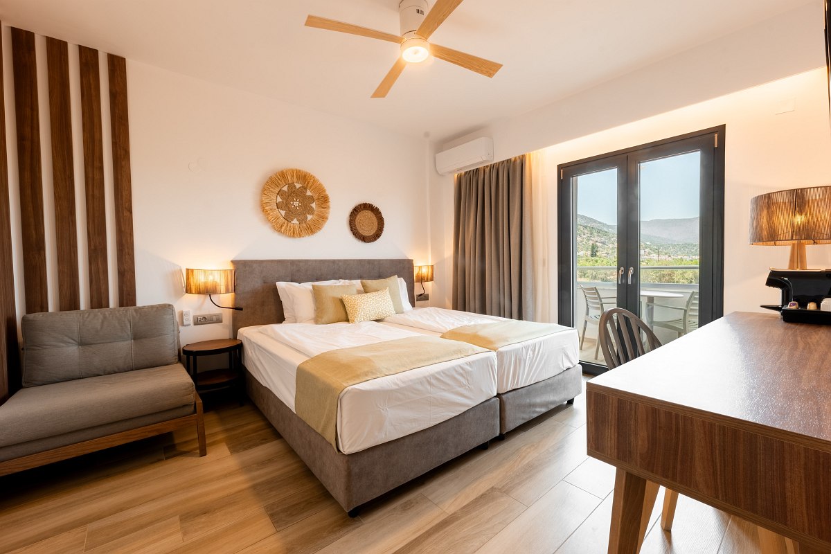 Modernes, helles Hotelzimmer mit Doppelbett, Sofa, Holzdekor und Balkon mit Blick auf Berge und Natur.