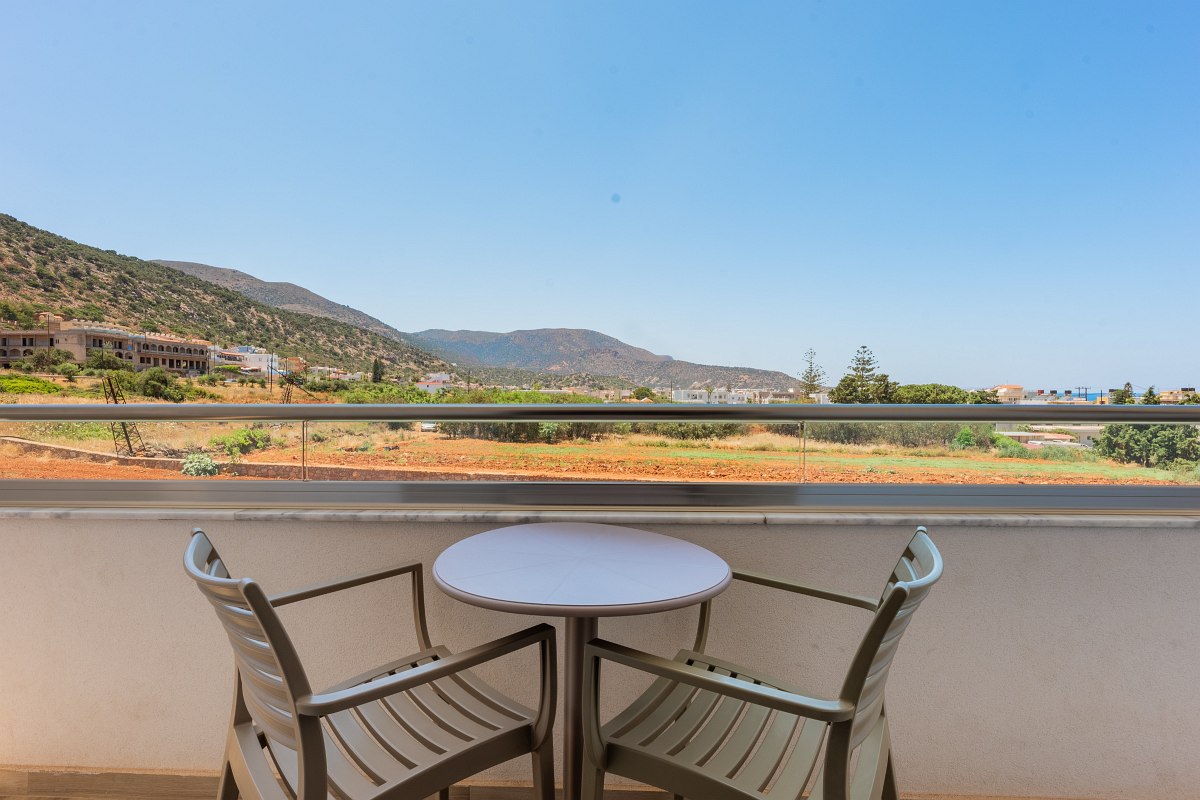 Balkon mit Tisch und zwei Stühlen, Blick auf grüne Landschaft, Felder und Berge unter klarem, blauem Himmel.