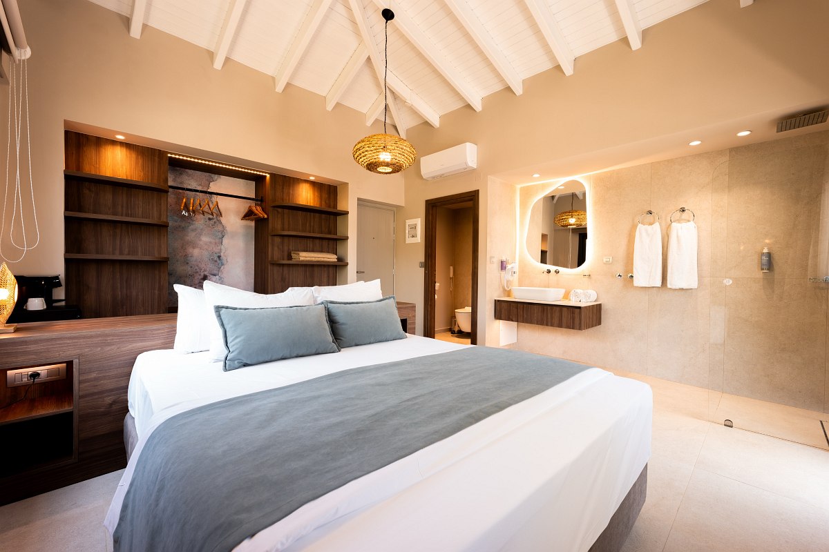 Helles Hotelzimmer mit gemütlichem Doppelbett, offenen Holzregalen, modernem Badbereich und freigelegten Dachbalken.