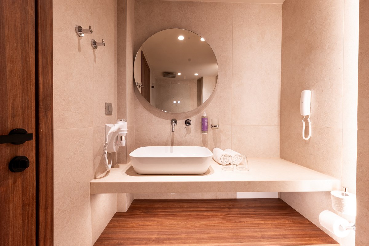 Modernes Hotelbad mit runder Spiegelleuchte, weißem Waschbecken, Holzdetails und großer begehbarer Dusche.
