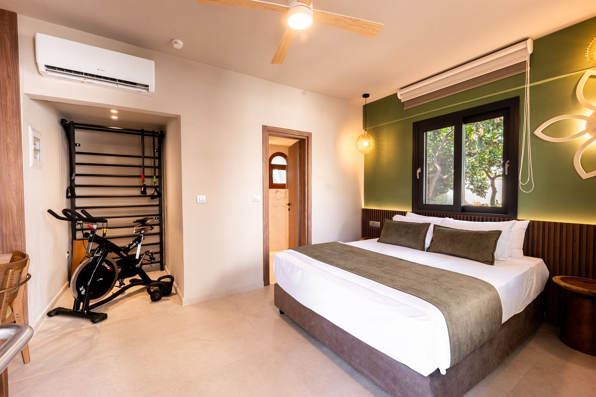 Modernes Hotelzimmer mit Doppelbett, Fitnessbereich, Spinning-Bike, grünem Akzent an der Wand und großem Fenster.