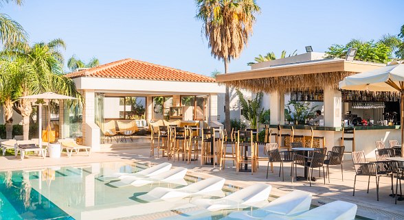 Sonnige Poollandschaft mit Liegen im Wasser, Bar mit Strohdach, Sitzbereich und Palmen im mediterranen Stil.