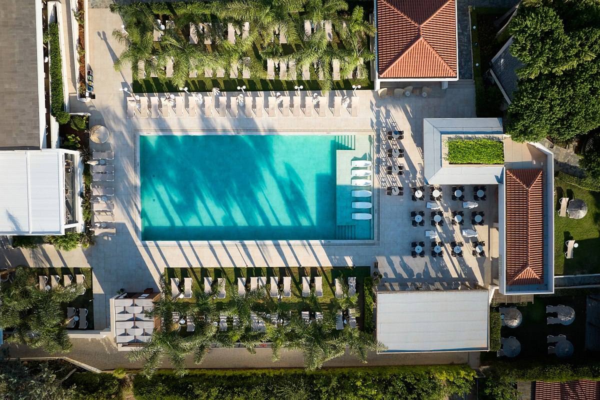 Großer Pool mit Liegen, Bar und Sitzbereichen unter Sonnenschirmen, umgeben von Palmen und mediterranen Gebäuden.