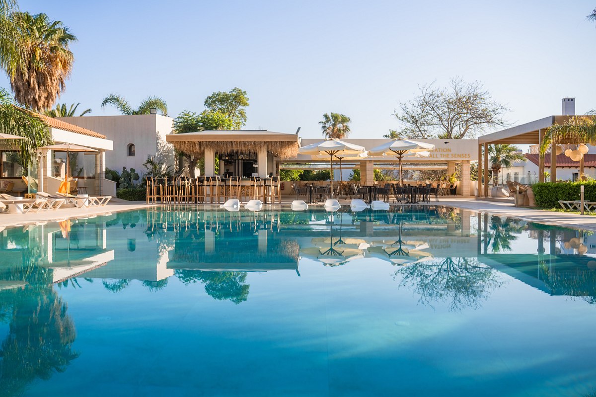 Großer Pool mit Liegen, Bar und Sitzbereichen unter Sonnenschirmen, umgeben von Palmen und mediterranen Gebäuden.