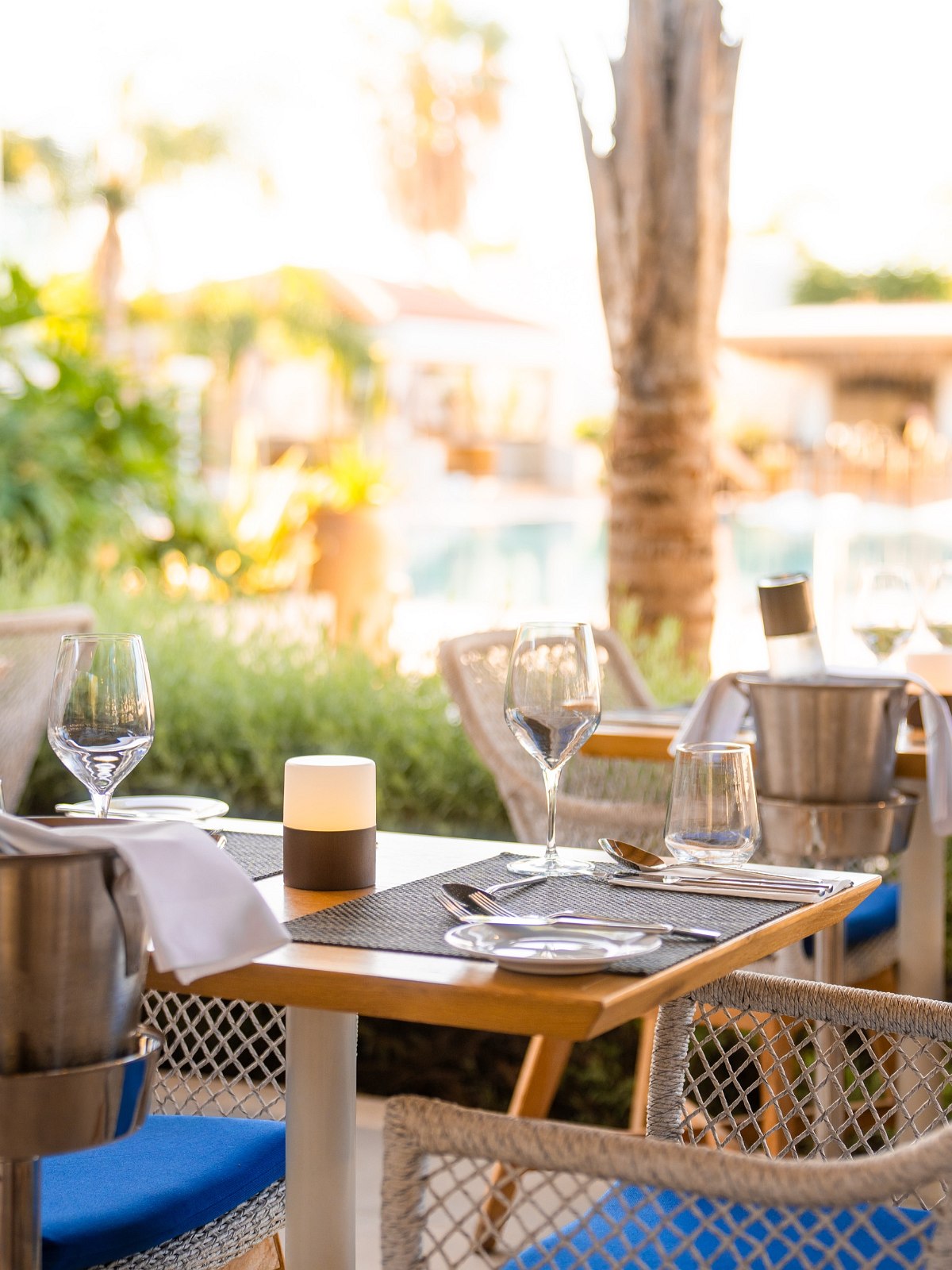 Elegant gedeckter Tisch im Außenbereich eines Restaurants mit Blick auf Palmen, Pool und Lounge-Bereich im Hintergrund.