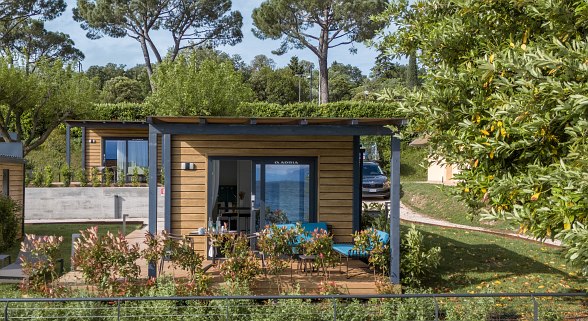 Modernes Mobilheim mit überdachter Terrasse, gemütlicher Sitzecke und Seeblick, umgeben von viel Grün und Bäumen.