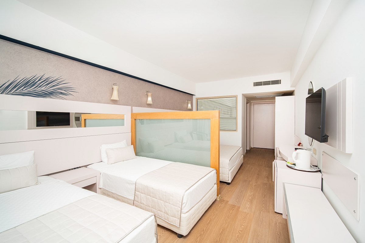 Modernes Hotelzimmer mit drei Einzelbetten, Raumteiler, TV, Schreibtisch und heller, schlichter Einrichtung.