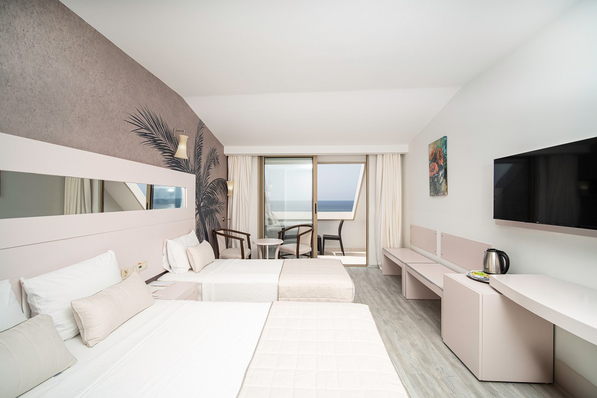 Modernes Hotelzimmer mit zwei Betten, Sitzbereich, Balkon mit Meerblick, Wasserkocher und TV an der Wand. 