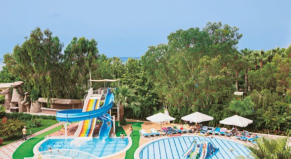 Poolanlage mit Wasserrutschen, Kinderbecken, Liegen und Sonnenschirmen, umgeben von grüner Gartenlandschaft.