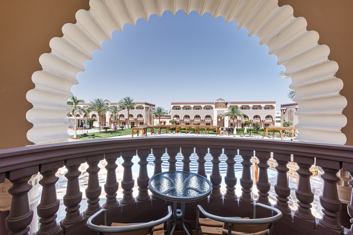 Balkon mit Glas-Tisch und Stühlen, eingerahmt von einem orientalischen Bogen, mit Blick auf Garten und Hotelanlage.